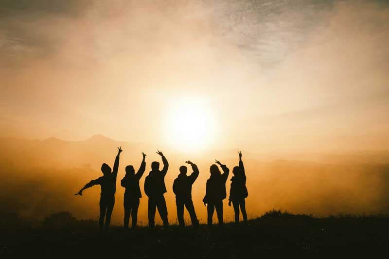 Silhouetted group of people on a hilltop with arms raised against a golden sunrise